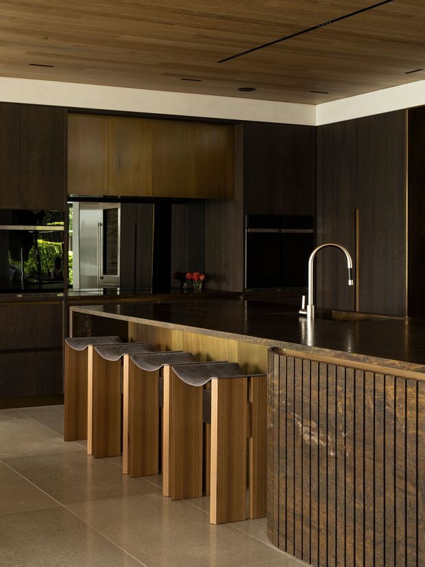 A sophisticated contemporary kitchen with dark timber cabinetry, a matching timber ceiling, and a large dark stone island. The island features a textured vertical timber-clad base and integrated seating for four with sculptural curved stools. A brushed metal gooseneck tap, integrated appliances, warm lighting, and polished neutral stone flooring complete the refined, moody interior.