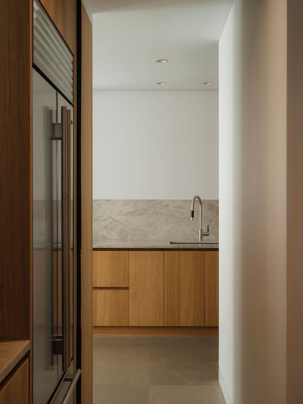 A minimalist kitchen viewed through a doorway, featuring warm timber cabinetry, a pale stone backsplash and benchtop, an undermount sink with a curved brushed-metal tap, recessed ceiling lights, and large neutral floor tiles. Dark timber and glass-framed joinery appear along the left side.