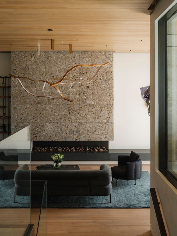 A refined contemporary living room viewed from an elevated angle, featuring a large textured natural-stone fireplace wall with a long linear gas fire, sculptural branching wall artwork, full-height glazing, and a warm timber-lined ceiling. A dark leather sofa and armchair sit on a muted blue-grey rug over timber flooring, with a glass balustrade in the foreground.