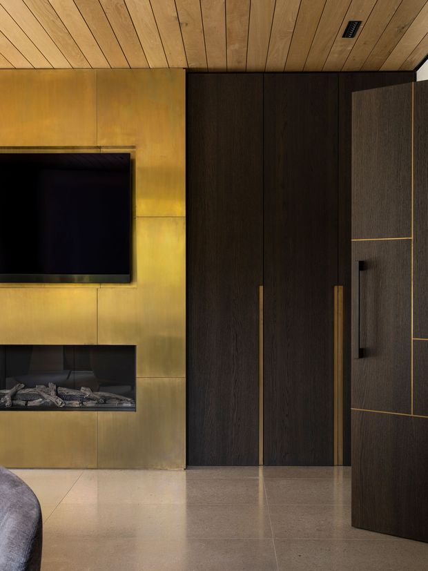 Close-up interior view of a refined media and storage wall. A large black television is mounted above a linear fireplace set within warm brass-toned metal wall panels. Adjacent floor-to-ceiling dark timber cabinetry provides concealed storage, with long brass handles and a darker textured panelled door. The ceiling is clad in warm timber boards, while the floor is finished in large polished light stone or concrete-look tiles. A partial upholstered sofa is visible in the foreground.