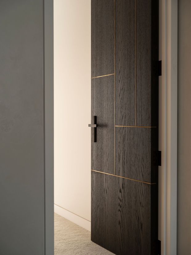 Close-up interior view of a partially open contemporary timber door with dark vertically grained wood veneer, slim brass inlay lines, and a dark metal lever handle. The door opens into a softly lit hallway or room with pale walls and carpet, framed by simple light-coloured trim.