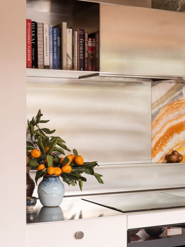 A close-up view of a refined contemporary kitchen with pale cabinetry, integrated open shelving, a reflective metallic backsplash, and a sleek induction cooktop above a built-in oven. The shelving displays cookbooks, while a marble or stone feature panel with warm orange veining appears at the right. A vase of citrus branches adds a natural decorative accent to the benchtop.