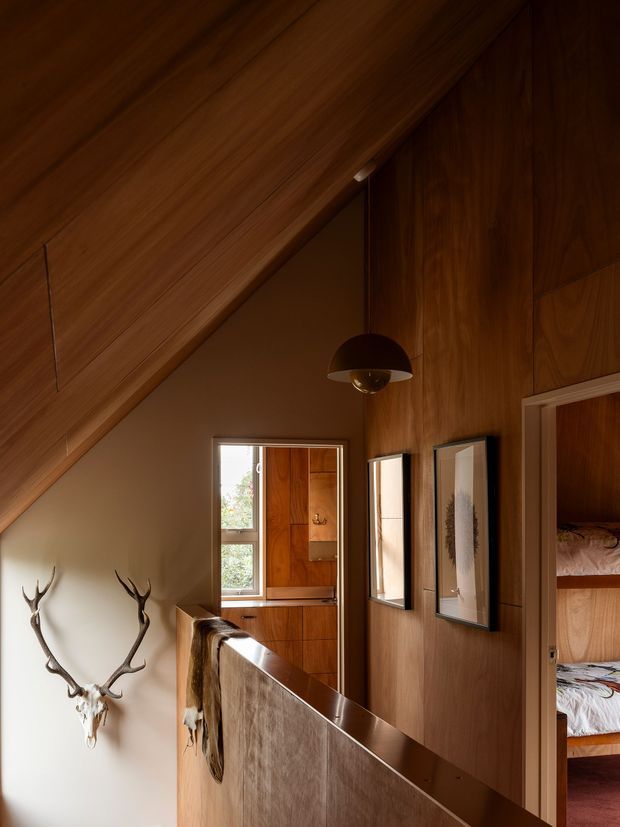 A warm, wood-lined upper-level hallway or landing beneath a steep sloping ceiling. The space features extensive dark timber wall and ceiling panelling, a half-height timber balustrade, framed artwork, a hanging dome pendant light, and decorative antlers mounted on a pale wall. An open doorway reveals a bedroom with bunk-style beds or layered bedding, while a window provides natural light at the end of the hall.