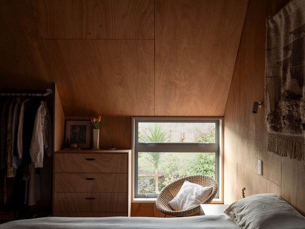 A warm, compact bedroom clad extensively in rich timber, with a sloping timber ceiling and matching wall panels. The room includes a low bed in the foreground, built-in timber drawers, an open clothes rail, a woven lounge chair, a framed picture, and a large horizontal window overlooking greenery. Soft natural light and earthy textures create an intimate, rustic atmosphere.