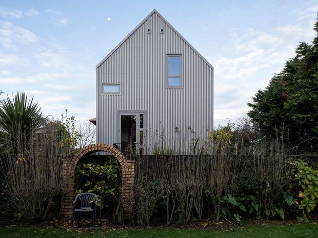 Front exterior view of a compact, narrow-gabled house clad in light grey vertical corrugated or profiled siding. The simple facade has a steep pitched gable roof, vertically arranged windows, a dark glazed entry door, and small circular ventilation openings near the gable peak. Dense garden planting, hedges, and a brick garden arch frame the foreground.