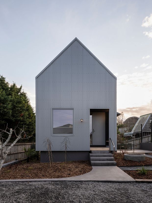 Front exterior view of a compact, narrow-gabled building with light grey vertical corrugated cladding, a steep pitched roof, one square window, and a recessed entry door reached by concrete steps. The structure sits in a landscaped residential setting with gravel paving, timber fencing, raised garden beds, and neighbouring structures visible at the sides.