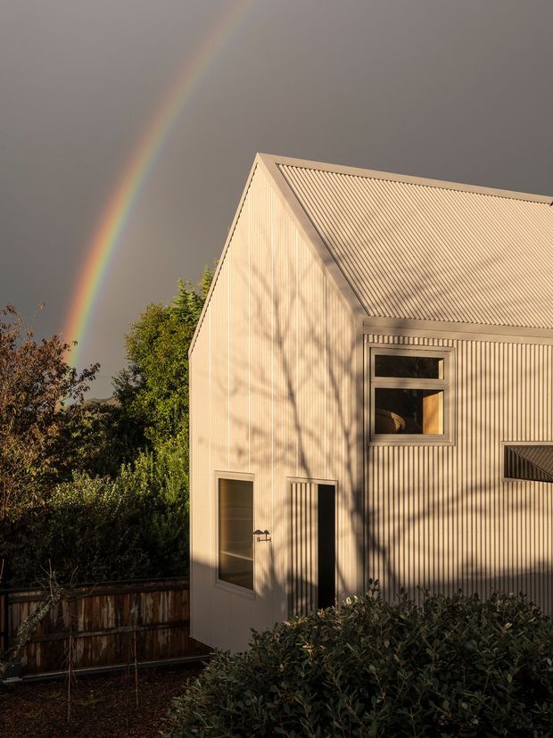 Exterior view of a compact gabled building clad in pale vertically ribbed weatherboard, with a corrugated pitched roof, simple rectangular windows and doors, and mature garden planting in the foreground. Warm low-angle sunlight casts tree shadows across the facade, with a rainbow visible against a dark sky.