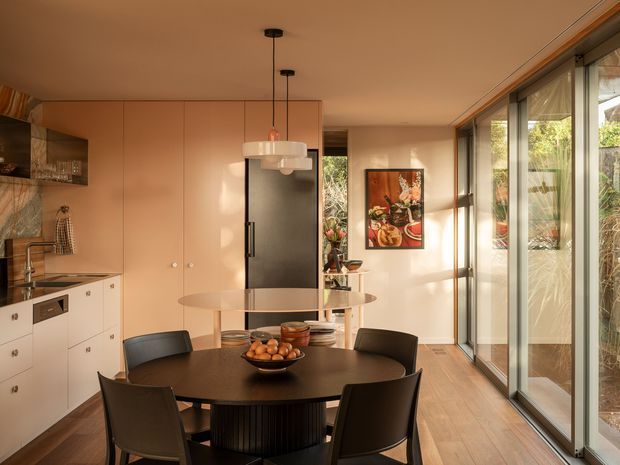 Warm contemporary open-plan kitchen and dining area with a compact dark dining table and four chairs, pale wood cabinetry, integrated dark refrigerator, stone-look benchtops, pendant lighting, and full-height glazed sliding doors opening toward a landscaped exterior. Timber flooring and peach-toned cabinetry create a cohesive mid-century-influenced interior.