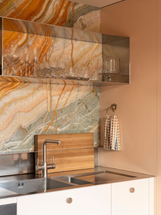 Close-up view of a refined compact kitchen or bar-style kitchenette. A dramatic orange, cream, grey and green-veined natural stone slab forms the backsplash and continues around the open display shelf. The space includes a stainless-steel sink and faucet, warm timber splashback detail behind the tap, pale cabinetry with round knobs, integrated open shelving displaying glassware, and warm peach-toned painted walls.