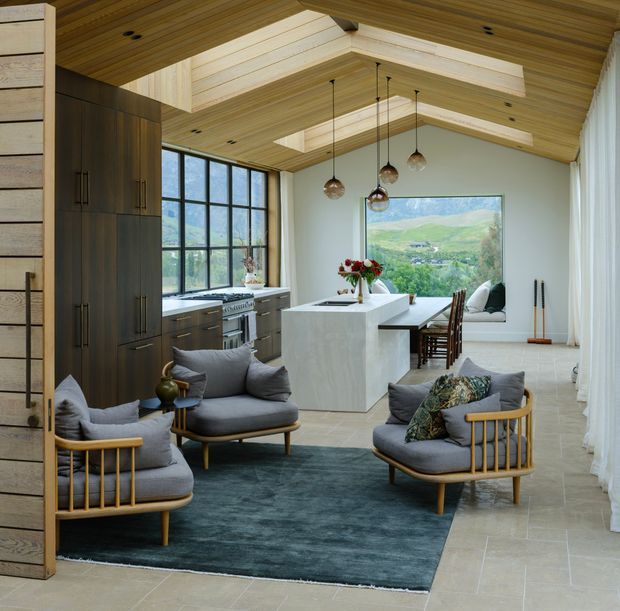 Open-plan kitchen and living area with a sculptural timber-lined ceiling, exposed roof framing, large black-framed glazing, dark timber cabinetry, a pale stone island, and a small seating arrangement around a deep blue rug. The room opens toward a scenic rural landscape through the windows and features warm pendant lights and neutral furnishings.