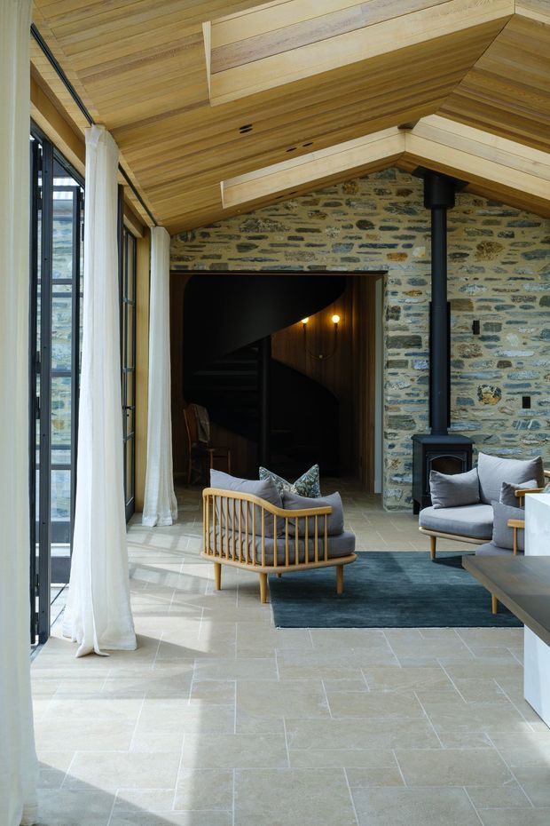 A refined open-plan living area with exposed stone masonry walls, a warm timber-lined vaulted ceiling, and expansive black-framed glazing with sheer curtains. The room includes a suspended-style black wood-burning stove, contemporary upholstered seating, a timber lounge chair, a dark area rug, and pale large-format stone or porcelain flooring. A dark stairwell and timber-lined interior are visible beyond the opening.