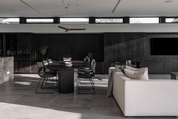 A sophisticated open-plan dining and living area with a dark monochromatic palette. A black dining table with matching contemporary chairs sits centrally, adjacent to a light upholstered sectional sofa. The space features dark timber or stone-look wall panelling, large-format grey floor tiles, a stone kitchen island, black-framed clerestory windows, recessed linear ceiling lighting, and a suspended ceiling fan. A television is integrated into the dark feature wall.
