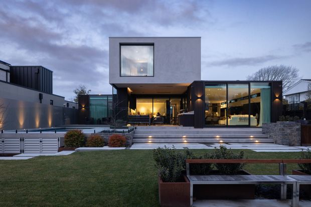 Twilight exterior view of a contemporary multi-level residence with a rectilinear rendered upper volume, extensive floor-to-ceiling glazing, dark framed openings, and warm interior lighting. Broad stepped terraces lead up from a landscaped lawn, with integrated garden seating, raised planters, low shrubs, and stone retaining walls.