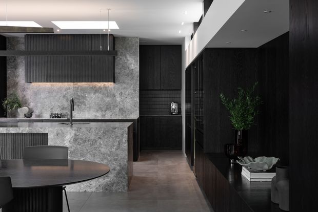 A sophisticated dark-toned contemporary kitchen and dining area. The space features a large grey stone island with integrated sink and black tapware, dark fluted and timber-look cabinetry, a textured stone splashback, black dining furniture, polished concrete-look flooring, and concealed linear lighting. A corridor-like view extends past full-height dark joinery and open shelving, creating a moody, refined interior.