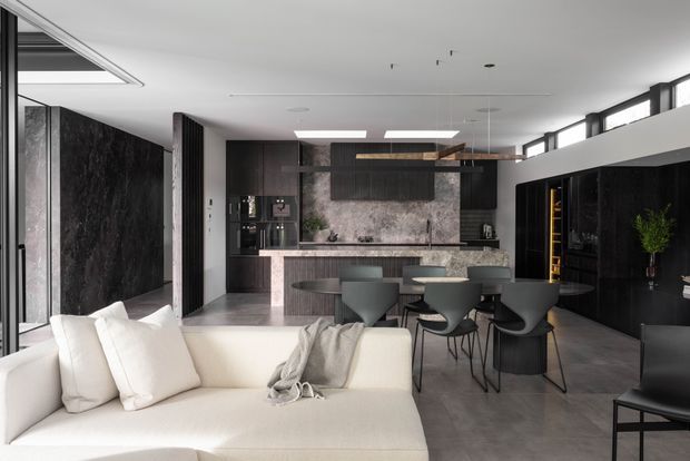 Open-plan contemporary living and kitchen interior with a white sectional sofa in the foreground, dark textured wall finishes and cabinetry, a central kitchen island, dining table with sculptural dark chairs, and extensive clerestory glazing. The space has a refined monochromatic palette with warm timber accents and polished concrete-like flooring.