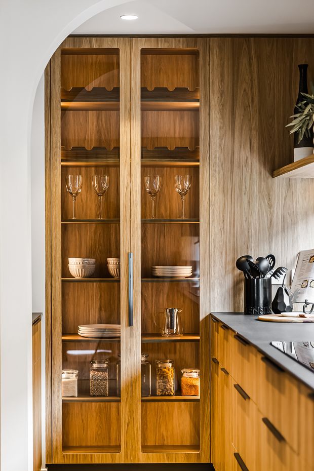 A warm, highly detailed kitchen storage area featuring a tall built-in timber display cabinet with glazed doors, open shelving, integrated lighting, glassware, crockery, and pantry jars. The cabinet sits beside timber kitchen cabinetry with dark benchtops and black hardware. A curved white plaster arch frames the left side, while a small section of open shelving and utensils is visible on the right.