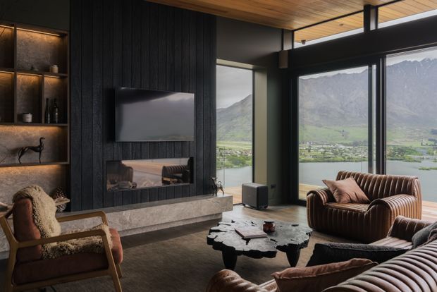 A moody, luxurious living room with floor-to-ceiling glazing overlooking a dramatic mountain and lakeside landscape. The room features a black vertical timber-panelled media wall with a built-in fireplace and shelving, a warm timber ceiling, dark-framed glazing, a sculptural coffee table, and richly upholstered brown leather seating.