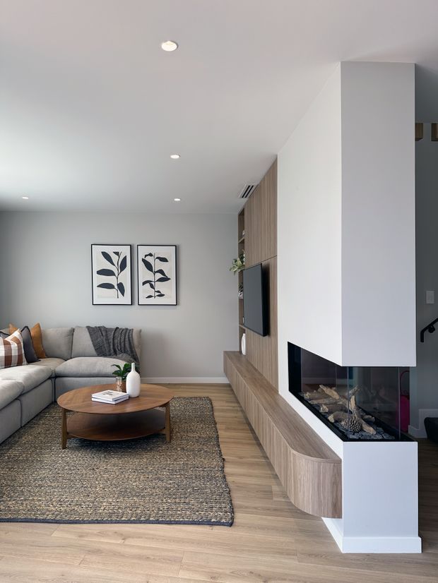 Contemporary living room with a large L-shaped grey sectional sofa, round timber coffee table, woven area rug, framed botanical artwork, and a built-in timber media wall. A sculptural white chimney breast incorporates a modern glass-fronted fireplace, with wood flooring and recessed ceiling lights creating a clean, warm interior.