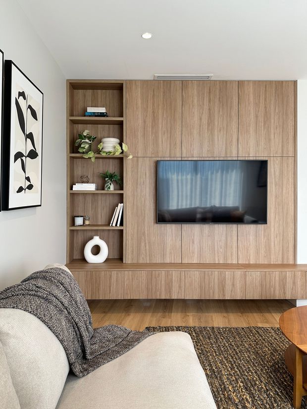 A warm, contemporary living room featuring a full-height timber entertainment wall with integrated television, open display shelving, and low cabinetry. Neutral upholstered seating, a textured dark rug, framed botanical artwork, indoor plants, and understated decor create a refined, inviting interior.