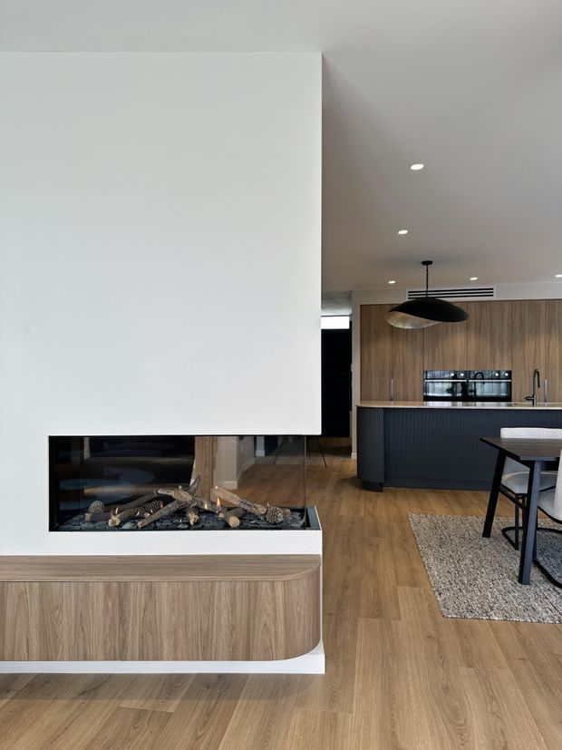 A contemporary open-plan living and dining area with a sculptural white fireplace feature in the foreground and a warm timber kitchen beyond. The room features timber flooring, vertical-grain timber cabinetry, a dark fluted kitchen island, integrated appliances, a black pendant light, recessed ceiling lights, and a dining table with upholstered chairs on a textured rug.