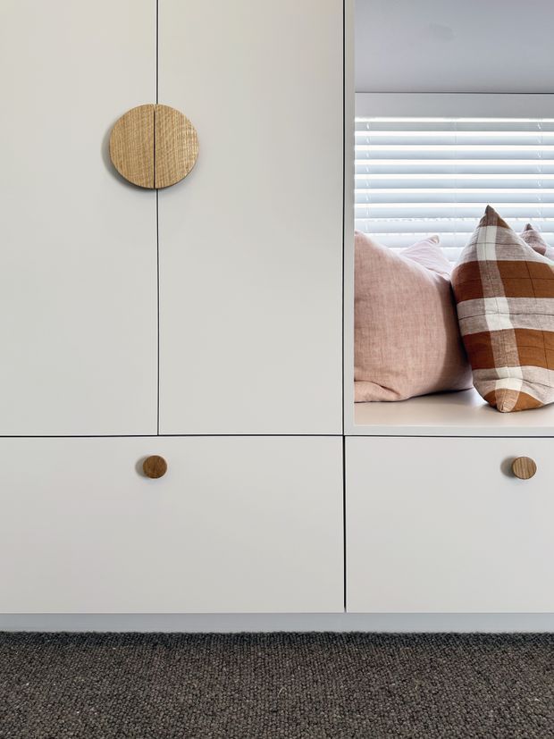 Close-up interior detail of a minimalist built-in bedroom storage unit in pale grey, with flush cabinet doors and small round timber knobs. An open cubby displays blush and brown checked cushions in front of white horizontal blinds. A dark textured carpet grounds the lower portion of the image.