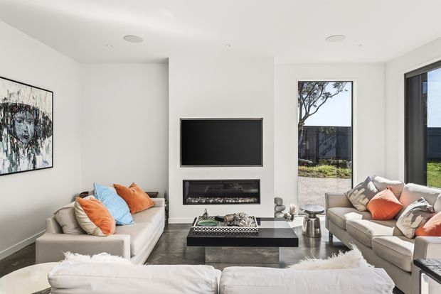Bright contemporary living room with white walls, a dark polished floor, built-in wall-mounted television, linear fireplace, central black coffee table, and two cream sofas arranged around the seating area. Large windows provide views of a landscaped exterior, while colourful cushions and abstract artwork add accents.