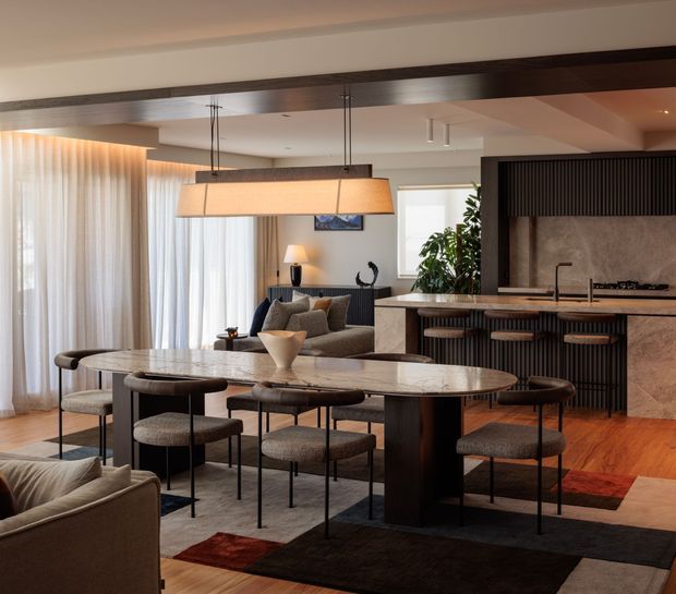 A sophisticated open-plan dining and kitchen interior with a large oval stone dining table, upholstered dining chairs, a dark timber structural beam, and a linear pendant light. The kitchen features dark vertically ribbed cabinetry, a stone island and splashback, timber flooring, and warm layered lighting. A living area is partially visible in the foreground, with sheer curtains and indoor planting adding softness.