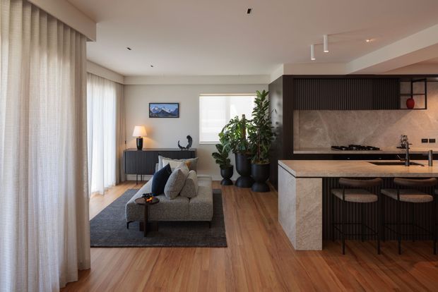 A refined open-plan living and kitchen area with warm timber flooring, a grey sofa and rug, full-height sheer curtains, indoor plants, and a dark detailed kitchen island. The kitchen features a stone-look benchtop and splashback, dark cabinetry or panelling, integrated lighting, and bar seating. Neutral finishes and abundant filtered daylight create a calm, contemporary interior.