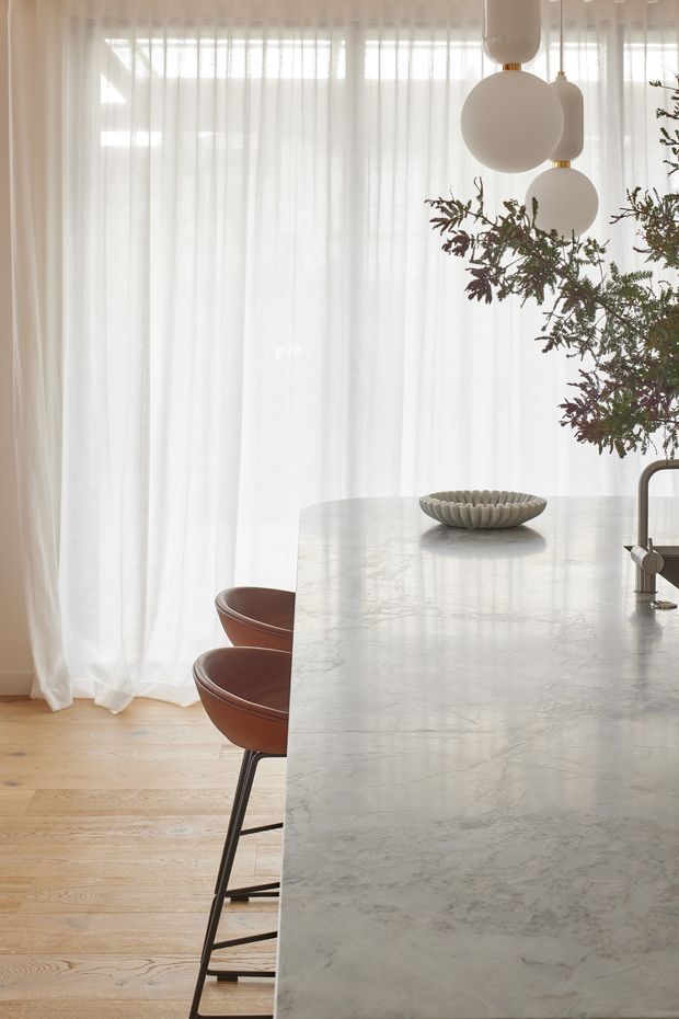 A softly lit contemporary kitchen interior focused on a large pale marble island with an integrated dark metal tap. Two curved brown leather bar stools sit along the island, while sheer white curtains cover tall glazing behind them. Sculptural white pendant lights hang overhead, and a leafy indoor branch arrangement adds organic contrast. The room has a warm, refined and airy atmosphere.
