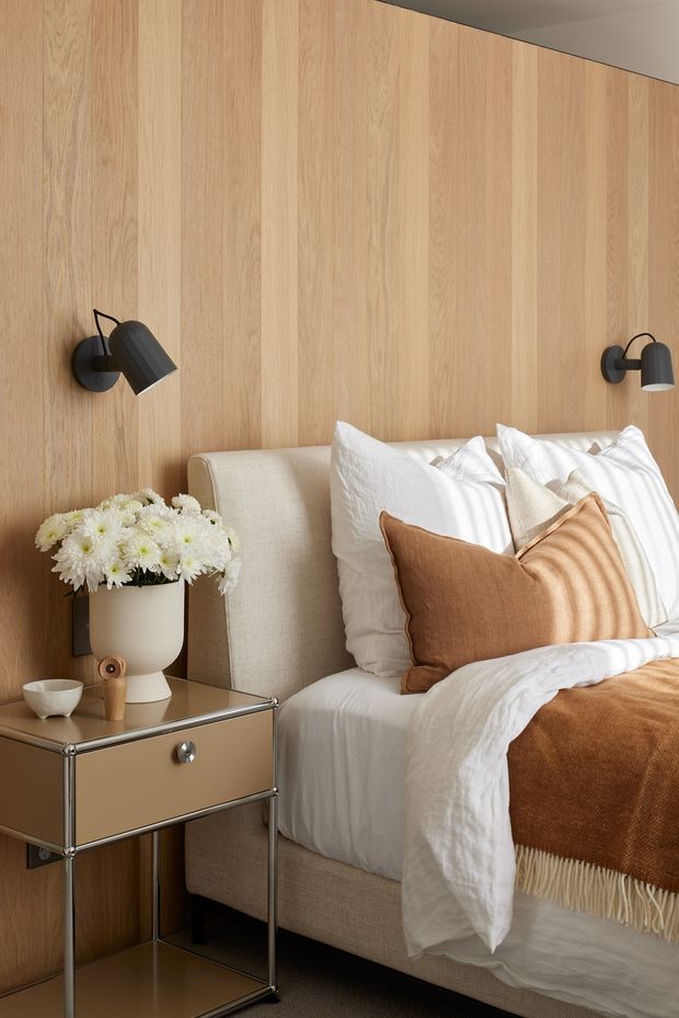 A warmly styled bedroom corner featuring a bed with layered white bedding, textured white pillows, and a rust-brown accent cushion and throw. The bed is set against a full-height light timber wall with vertical panel joints. Two dark wall-mounted reading sconces flank the bed, and a compact metallic bedside table holds a vase of white flowers and small ceramic and wooden objects. The palette is warm, neutral, and softly textured.