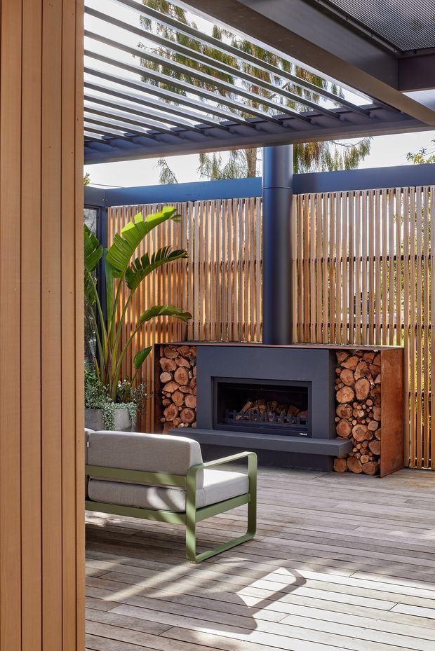 A covered outdoor entertaining area viewed from an interior doorway, featuring a built-in dark fireplace with integrated firewood storage, timber slat privacy screening, a timber deck, a black metal pergola with translucent roofing or slatted overhead structure, and a lounge chair surrounded by tropical planting.
