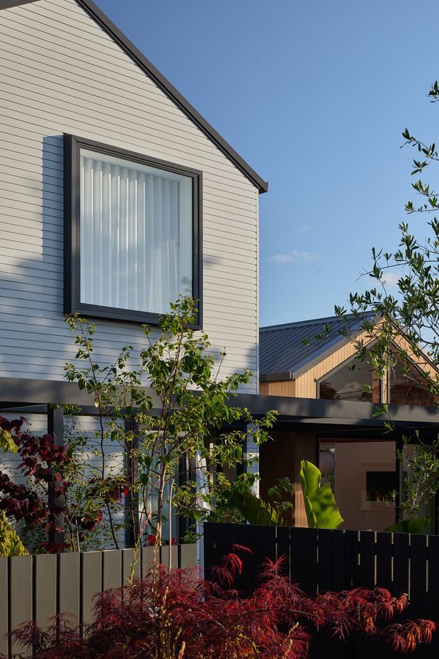 Exterior view of a contemporary multi-level residence with light horizontal weatherboard cladding, dark metal roof edging, large dark-framed window, and a covered lower-level outdoor area. Layered planting and dark privacy fencing occupy the foreground, with a second gabled wing visible behind.