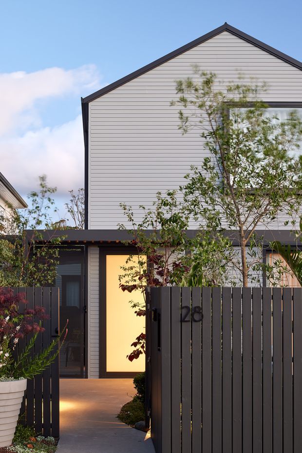 Exterior view of a contemporary multi-storey townhouse, showing a steep gabled roof, pale horizontal cladding, dark trim, and a recessed entry behind a tall dark vertical-slat fence. Warm light glows from the front door and entryway, with established landscaping in the foreground.