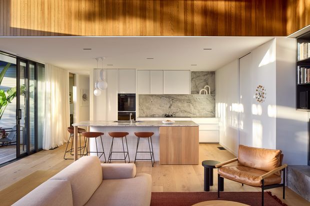 An open-plan contemporary kitchen and living area with a warm timber-lined mezzanine or upper wall, white full-height cabinetry, a large natural-stone island splashback, and timber flooring. The kitchen includes a breakfast bar with three stools, integrated appliances, and pendant lights. The living zone has a light upholstered sofa, a leather lounge chair, a black side table, and a low-profile rug. Full-height glazing and sheer curtains bring in abundant sunlight, while open shelving and a television are visible at the right.