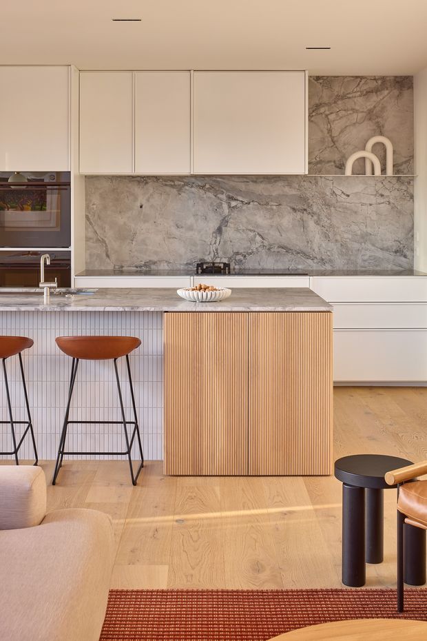 A refined contemporary kitchen viewed from the living area, featuring a large island with vertically fluted timber cabinetry, a pale stone benchtop and full-height veined stone splashback. The kitchen has soft white overhead and base cabinetry, integrated appliances, a linear tiled island face, timber flooring, and warm sculptural bar stools. The palette is warm and neutral with terracotta and dark accents, creating a polished yet inviting interior.