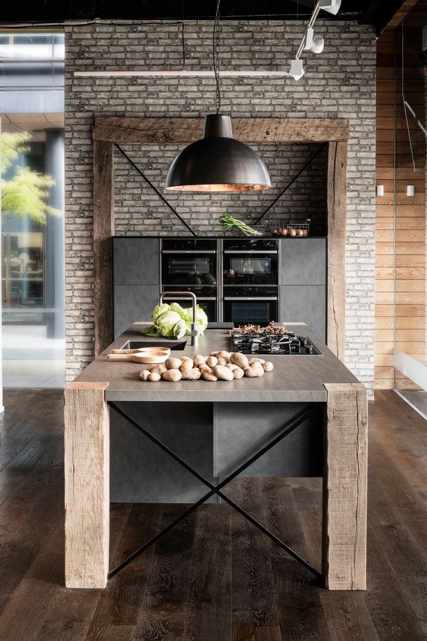 Industrial-style kitchen centred on a large island with a dark stone-look benchtop, integrated sink and cooktop, black cabinetry, and timber framing. A weathered brick wall forms the backdrop, with a large black dome pendant light, exposed dark ceiling, timber wall cladding, and dark timber flooring.