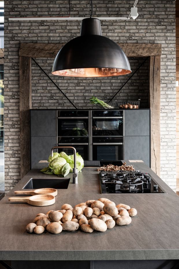 A dramatic industrial-style kitchen with a large dark stone island, integrated black sink and gas cooktop, dark cabinetry, twin ovens, exposed grey brick walls, and rustic timber framing. A large black pendant light with a copper-toned interior hangs above the island, creating a warm focal point.