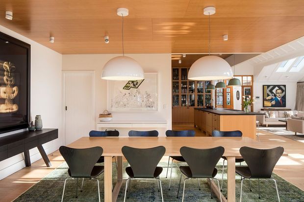 Open-plan dining and kitchen interior with a long light-wood dining table, sculptural dark chairs, oversized white pendant lights, and a warm timber-lined ceiling. A timber kitchen with island and bar seating is visible behind the dining area, along with built-in shelving, artwork, and abundant daylight from roof glazing. The room has a refined mid-century-inspired character with warm natural materials and strong indoor-outdoor light.