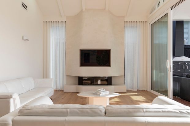 Bright, refined living room with white upholstered sofas arranged around a low sculptural coffee table and a built-in fireplace feature wall. Tall sheer-draped windows and sliding glass doors provide abundant natural light, with a glimpse of an outdoor entertaining area beyond.