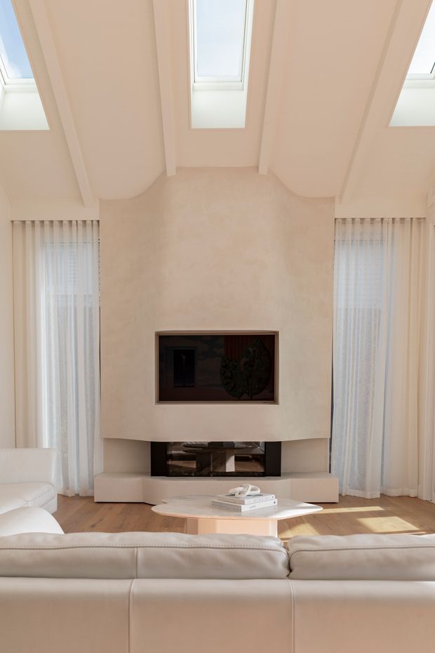 Bright, refined living room centred on a sculptural plaster fireplace wall with a dark recessed firebox and lower open hearth. White leather sofas face the fireplace, with a low rounded coffee table on warm timber flooring. Tall sheer-curtained windows flank the fireplace, while multiple roof skylights and expressed ceiling beams bring in abundant natural light.