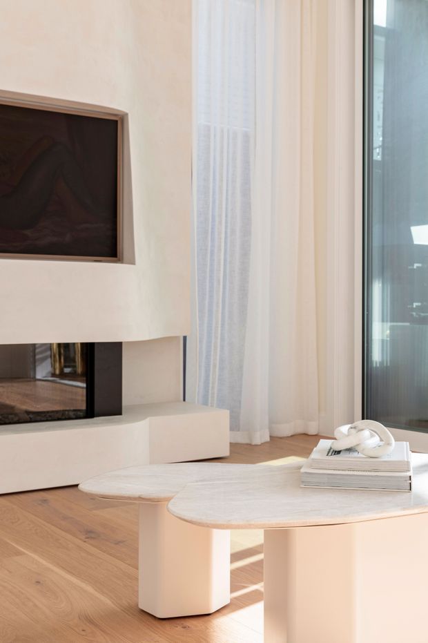 Bright contemporary living room with a minimalist built-in fireplace and television wall, sheer floor-to-ceiling curtains, large glazed opening, pale timber flooring, and sculptural rounded coffee tables in a warm neutral palette.