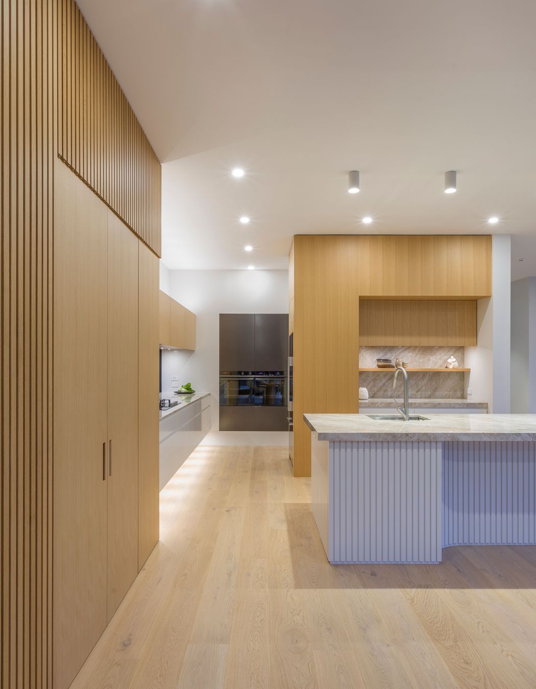 Weaving materiality with form, oak veneer warms the tall walls with battens expressed in an L-shaped motif.  This motif is echoed in veneer treatments of both tall walls and subtly evident in the island plan.