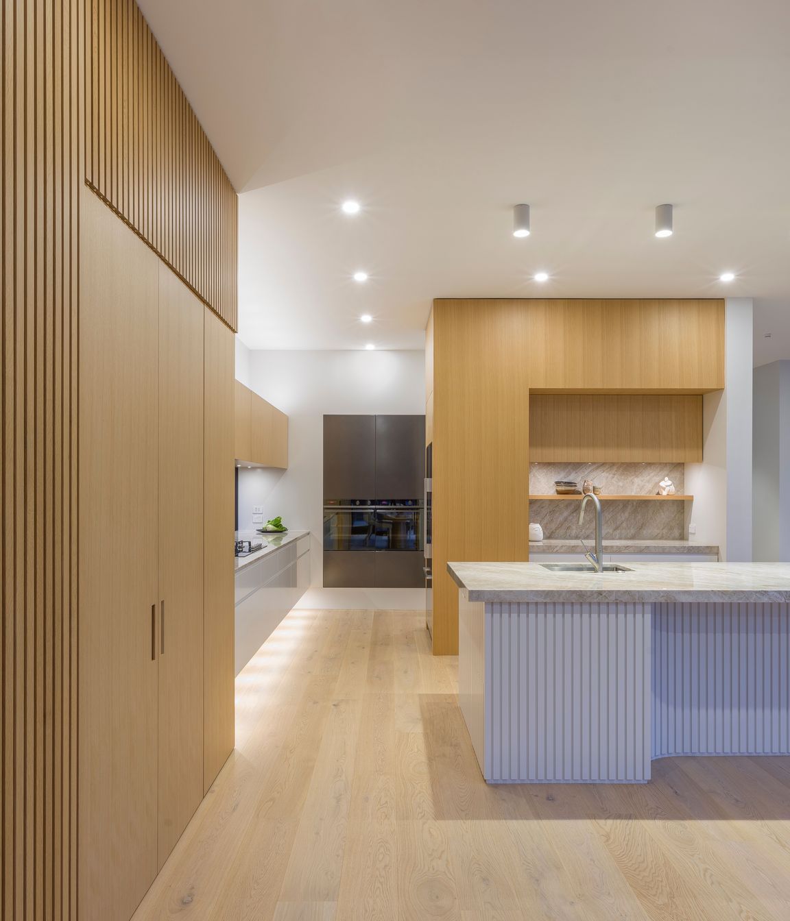 Weaving materiality with form, oak veneer warms the tall walls with battens expressed in an L-shaped motif.  This motif is echoed in veneer treatments of both tall walls and subtly evident in the island plan.