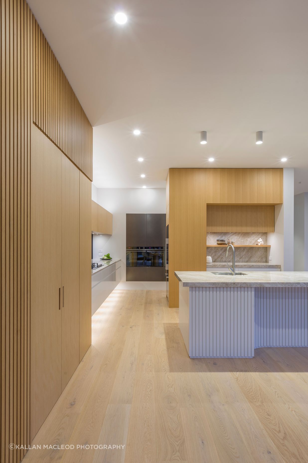 Weaving materiality with form, oak veneer warms the tall walls with battens expressed in an L-shaped motif.  This motif is echoed in veneer treatments of both tall walls and subtly evident in the island plan.