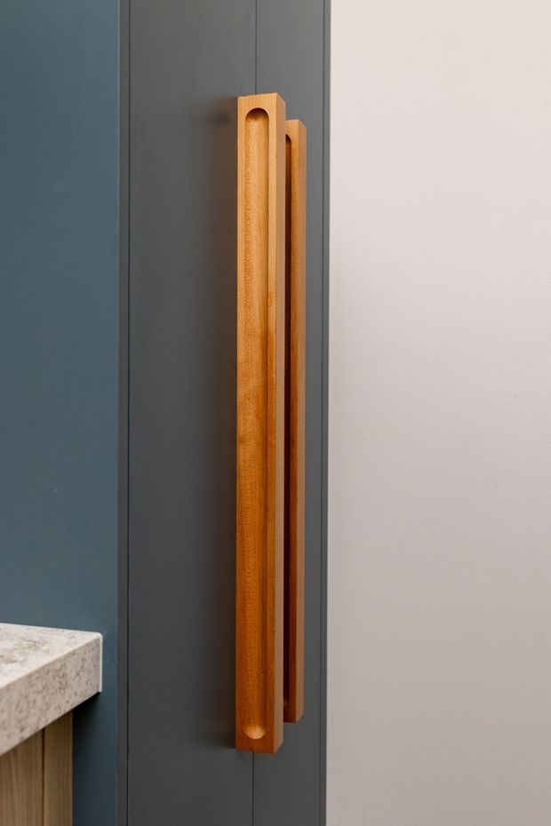 Close-up detail of a tall vertical solid timber pull handle mounted on dark charcoal cabinetry or a door. The handle has a warm orange-brown wood finish with a recessed rounded channel, contrasting against the dark grey joinery and adjacent light neutral wall. A small portion of a pale stone benchtop and timber joinery is visible at the lower left.