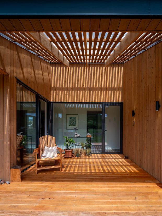 A warm timber-lined courtyard or covered outdoor terrace framed by vertical wood walls and a slatted pergola roof. Sunlight creates strong linear shadows across the timber decking and rear glazing. Black-framed glass doors open toward an interior space, while a wooden lounge chair, potted plants, and minimal furnishings add a relaxed, natural character.