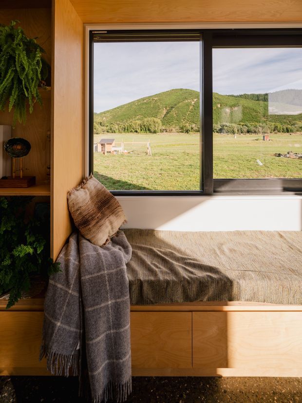A warm, compact built-in window seat in a timber-lined interior, with integrated shelving and cabinetry beneath a large black-framed window. A textured beige cushion and grey checked throw create a cosy reading nook, while the window overlooks an open rural valley and green hills. The space is softly lit by sunlight and uses natural timber, neutral textiles, and dark speckled flooring.
