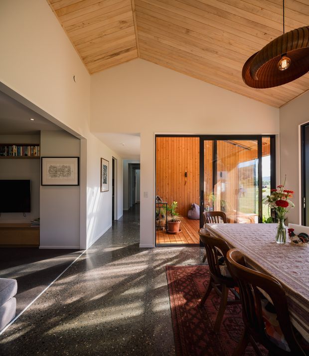 A warm, light-filled open-plan dining and living area with a high pitched ceiling lined in timber boards. The room features polished dark flooring, a large dining table with dark wood chairs, a black woven pendant light, built-in media shelving, and framed artwork. Full-height glazed doors open onto a timber-lined deck or sunroom, bringing in views and abundant natural light.