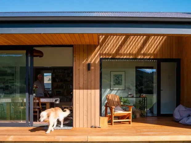 Single-storey modern home viewed from a timber deck, with warm vertical timber cladding, a broad low-pitched metal roof, extensive dark-framed glazing and sliding doors. The open interior includes a dining or living area, while the deck has timber seating and a relaxed outdoor living arrangement. Strong sunlight creates patterned shadows across the facade and deck.