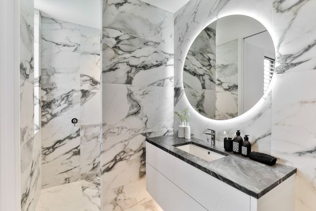 Contemporary bathroom featuring dramatic white and grey veined marble-look wall and floor surfaces, a floating white vanity with dark stone benchtop, integrated basin, chrome tapware, and a large round backlit mirror. A glass-enclosed shower is visible at left.